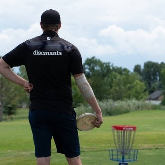 Discmania sponsored disc golf player Simon Lizotte standing back to holding a disc against his leg wearing an elbow brace.