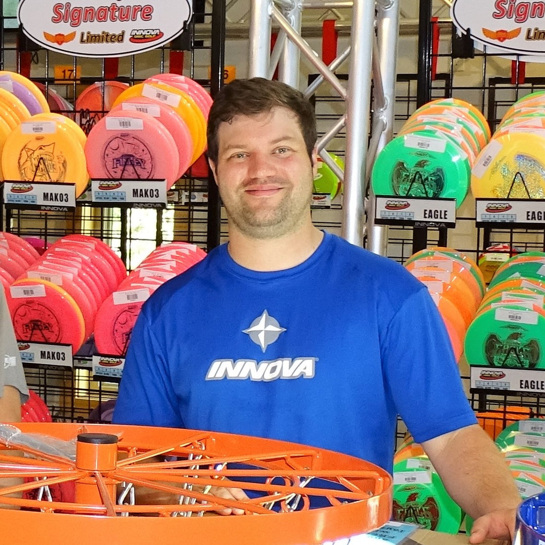 Sabattus Disc Golf blog writer and Pro Shop staff, Andrew Streeter, standing in front of a full rack of colorful discs.