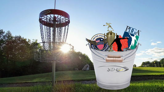 A bucket full of disc golf related items next to a disc golf basket with the setting sun behind it.