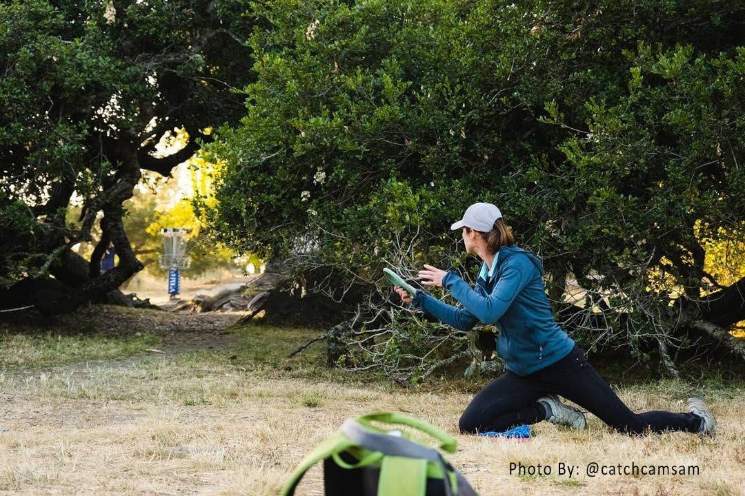 A female disc golfer leans out from behind bushes kneeling, to make an upshot to a basket in a gap.