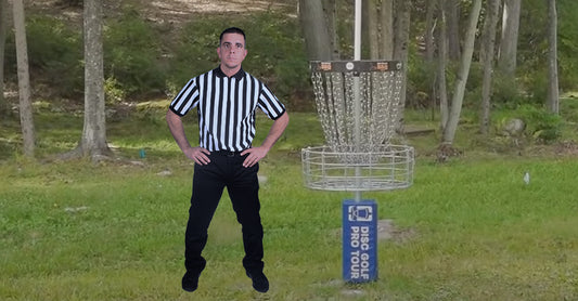 Black & white Striped Shirted Referee standing next to a disc golf basket