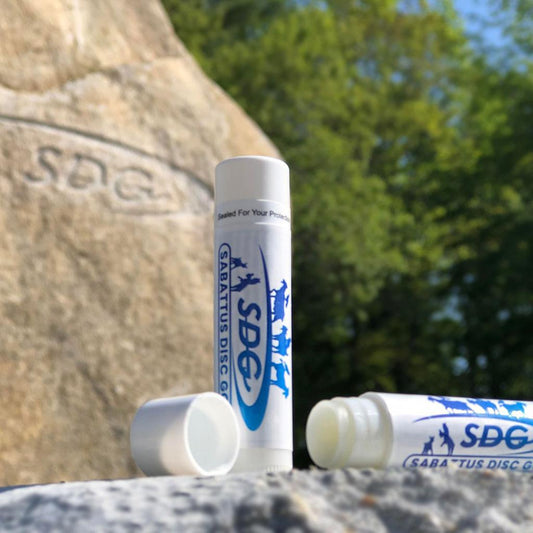 Sabattus Disc Golf branded original chapstick outside in front of SDG engraved rock, showing Blue SDG swish with goats and birds and the Sabattus Disc Gold Bar logo on two tubes of chapstick one open with cover off to the side the other straight up and down 