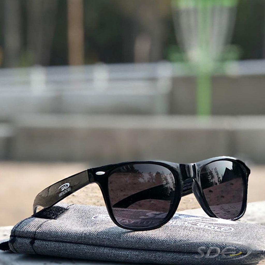 Sabattus Disc Golf Branded black sunglasses with Sabattus Disc Golf Branded Heathered Gray Sunglasses Sleeve outside with green disc golf basket in background 